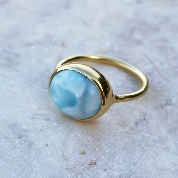 A gold ring featuring a smooth, oval-shaped light blue larimar stone, photographed on a textured light gray stone surface.