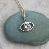 Silver eye-shaped pendant with an aquamarine gemstone on a chain, placed on a green stone.