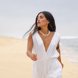 Woman in a white jumpsuit standing on sand with a blurred background wearing white beaded shell necklace with larmar and larimar eye pendant on golden chain
