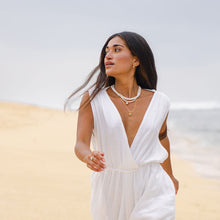 Load image into Gallery viewer, Woman in a white jumpsuit standing on sand with a blurred background wearing white beaded shell necklace with larmar and larimar eye pendant on golden chain
