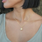 A woman with short black hair bob wearing a delicate gold necklace with citrine pendant in gold and matching gold hoop earring with round citrine gemstones, photographed outdoors against a textured stone background.