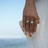 A close-up of a person's hand resting on a textured white surface, wearing two gold rings with light blue Larimar gemstones. The background is a soft, out-of-focus light blue and beige scene.