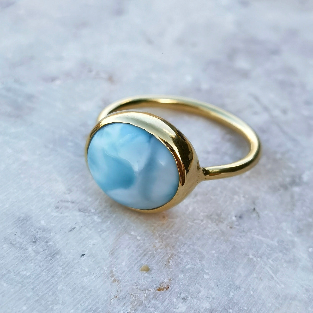 A gold ring featuring a smooth, oval-shaped light blue larimar stone, photographed on a textured light gray stone surface.