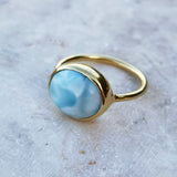 A gold ring featuring a smooth, oval-shaped light blue larimar stone, photographed on a textured light gray stone surface.