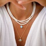 Close-up of a person wearing white outfit and multiple white shell beaded necklaces with larimar stones set in 18ct gold plated silver and a larimar eye necklace in gold