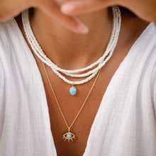 Load image into Gallery viewer, Close-up of a person wearing white outfit and multiple white shell beaded necklaces with larimar stones set in 18ct gold plated silver and a larimar eye necklace in gold
